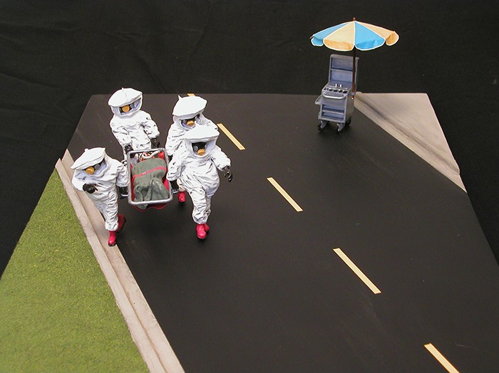 A diorama of 4 people in hazmat suits, carrying a person on a gurney with a hotdog stand in the background – created by Rupert Nesbitt. 14”x 30”x 6”, Sculpy, Styrene, 2004